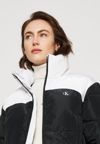 Giacca a vento bianca e nera con collo alto, caratterizzata da una texture trapuntata, chiusura con zip e logo CK sul petto.
