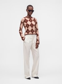 Personne portant un pull à losanges marron et beige, un pantalon blanc taille haute, des chaussures noires pointues et des lunettes de soleil noires, debout avec une main dans la poche.