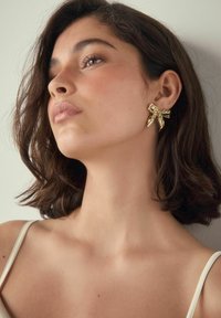 Goldfarbener, schleifenförmiger Ohrring mit filigranen Details. Am Ohr befestigt, mit glatter, glänzender Textur und einem mehrlagigen Banddesign.