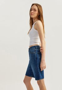 Bermudas de mezclilla en azul oscuro con cintura ajustada, textura suave, diseño de cinco bolsillos y detalles de dobladillo sin acabar. Combinadas con una camiseta sin mangas blanca.