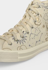Μπεζ sneaker ψηλού τύπου με μπεζ κορδόνια και ασημένιες και χρυσές εικονογραφήσεις με θέμα τα ουράνια σώματα, όπως αστέρια, σύννεφα και ο ήλιος.