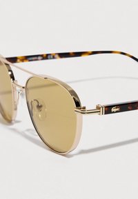 Okulary przeciwsłoneczne z złotą metalową oprawką, zielonkawo-brązowymi soczewkami, zausznikami w wzór żółwia oraz logo przy zawiasie.