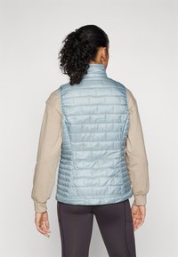 Lys blå dunvest med høy krage, med et kviltet mønster og en tettsittende design. Bæres over en tan langermet skjorte.