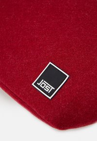 Tissu rouge texturé avec une étiquette rectangulaire de la marque JOST en noir et blanc cousue près du bord.