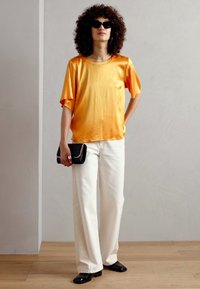 Top giallo in satin a maniche corte, pantaloni bianchi a gamba larga, borsa nera con catena, occhiali da sole neri, capelli ricci, in piedi davanti a uno sfondo neutro.