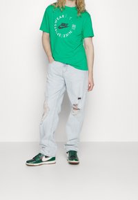 Vihreä Nike t-paita pyöreällä graafisella logolla, vaaleansiniset revityt farkut ja vihreät Nike lenkkarit valkoisilla yksityiskohdilla ja ruskeilla pohjilla.