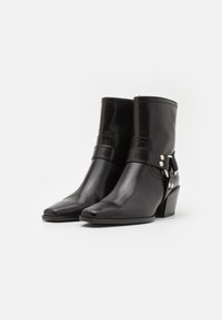Bottines en cuir noir avec un bout pointu, un talon bloc de hauteur moyenne et des accents de sangle avec quincaillerie argentée. Texture lisse.