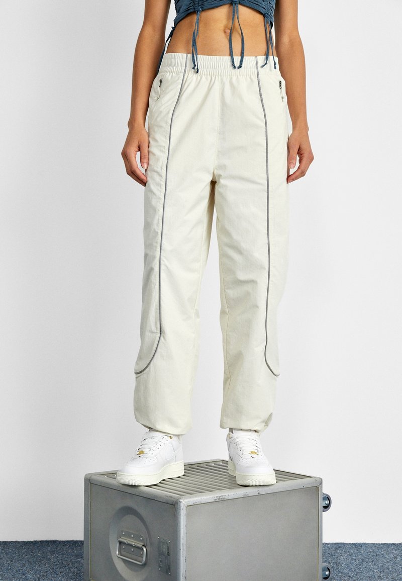 Lätta, krämfärgade cargobyxor med elastisk midja, sidofickor och grå detaljer. Bärs med vita sneakers på en metallkruka.