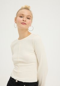 Ribgebreide top met lange mouwen in roomwit, met een ronde halslijn en contrasterende stiksels langs het midden. Gecombineerd met zwarte broek en ronde oorbellen.