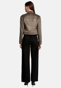 Veste courte en daim brun avec un col haut, des détails au dos, et des manches longues, associée à un pantalon noir à jambes larges.