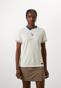 Biely dres s krátkym rukávom a modrým golierom, s logom kohúta a značkou Nike. Textúrovaná tkanina s červenými a modrými akcentmi na rukávoch.