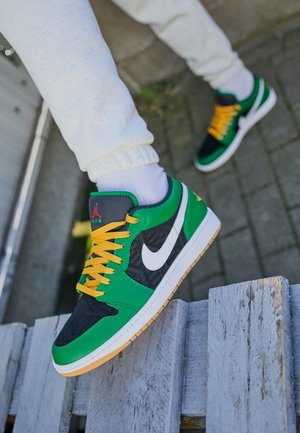 Osoba nosząca zielone, czarne i białe buty Nike Air Jordan z żółtymi sznurowadłami, stąpająca po nadwyrężonej drewnianej powierzchni na zewnątrz.