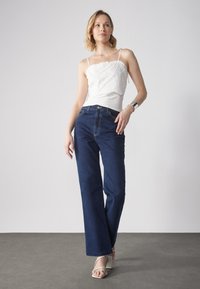 Hvit ermeløs topp med blomsterbroderi, kombinert med mørkeblå vide jeans. Modellen har på seg sølvtilbehør og hvite hæl-sandaler.