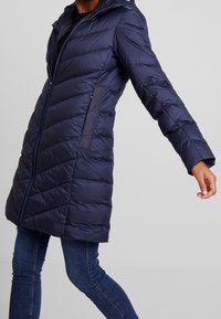 Mörkblå puffjacka med quiltad design, dragkedja fram, huva och sidofickor. Tillverkad av lätt, isolerat material med en slät yta.