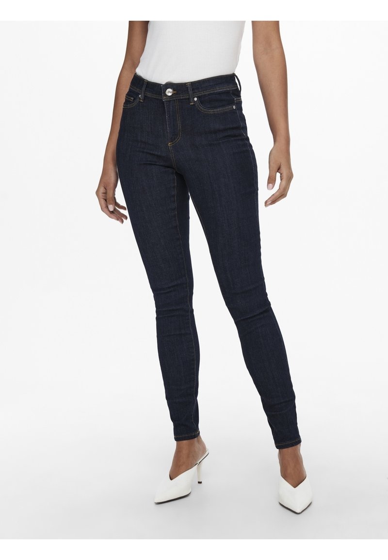 Mujer vestida con jeans ajustados de color azul oscuro, top sin mangas blanco y mules de tacón puntiagudos blancos, de pie contra un fondo blanco.