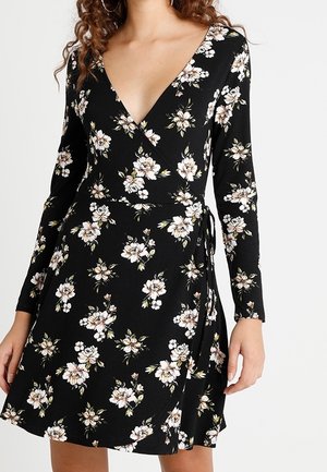 Black long-sleeve wrap dress with white and beige floral pattern, tied at the waist, worn by a woman with curly hair.