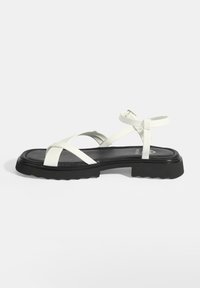 Witte leren sandalen met een zwarte rubberen zool, voorzien van gekruiste banden en een enkelgesp. Plat ontwerp met een kleine plateau.