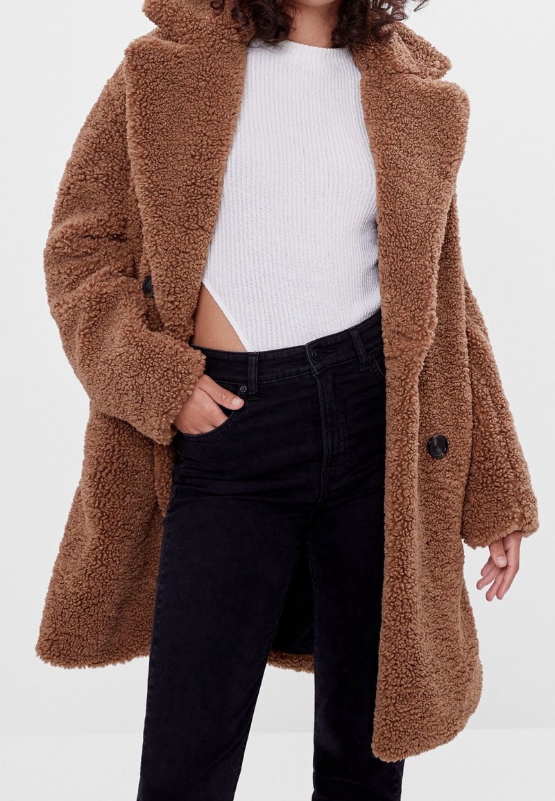 Bruine, zachte sherpa-stijl jas met een gekarteld kraag, twee knopen en zijzakken, gecombineerd met een witte ribbeltop en zwarte jeans.