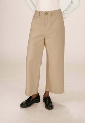 Personne portant un pantalon cropped beige à jambes larges avec des poches avant et des mocassins noirs, debout sur un fond clair.