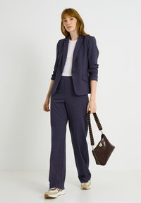 Traje azul marino con estampado, que incluye un blazer entallado y pantalones de pierna recta, combinado con un bolso marrón con detalles de lunares y zapatillas blancas.