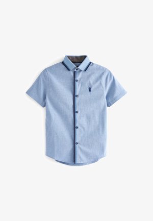 Chemise à manches courtes bleu clair avec bordure bleu foncé sur le col et la ligne de boutons, avec un petit logo de cerf bleu foncé sur la poitrine.