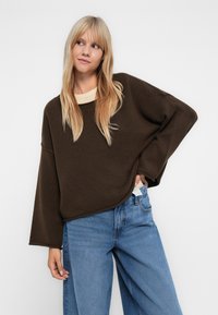 Suéter oversized castanho escuro com decote redondo e ombros caídos, combinado com jeans de perna larga azul claro. Textura suave, ajuste casual.