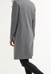 Cappotto di lana grigio con un design su misura, maniche lunghe e polsini abbottonati. Presenta una silhouette elegante e arriva a metà coscia su chi lo indossa.