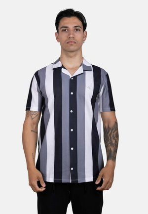 Joven con cabello corto y oscuro que lleva una camisa de manga corta a rayas verticales en negro, blanco y gris, de botones, de pie frente a un fondo liso.