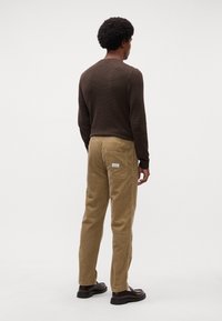 Chemise à manches longues texturée marron associée à un pantalon en velours côtelé beige avec une poche arrière, portée avec des chaussures noires, vue de dos.