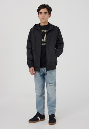 Giacca leggera nera con cappuccio e chiusura con zip, abbinata a una t-shirt grafica nera e a jeans azzurri chiari strappati, e sneakers nere.