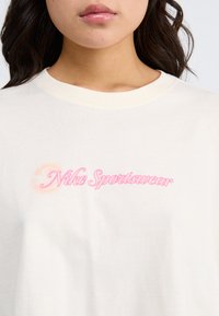 Krämfärgad bomullströja med rund halsringning och korta ärmar, med en rosa "Nike Sportswear"-logotyp på bröstet. Mjuk textur.