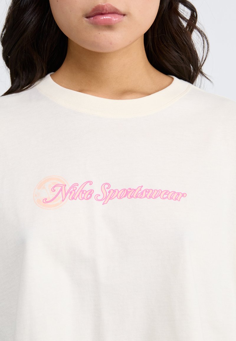 Camiseta de algodón color crema con cuello redondo y mangas cortas, que presenta un logo rosa de "Nike Sportswear" en el pecho. Textura suave.