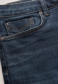Primo piano di jeans in denim blu scuro che mostra la tasca porta monete e la tasca frontale con cuciture marroni e rivetti metallici.