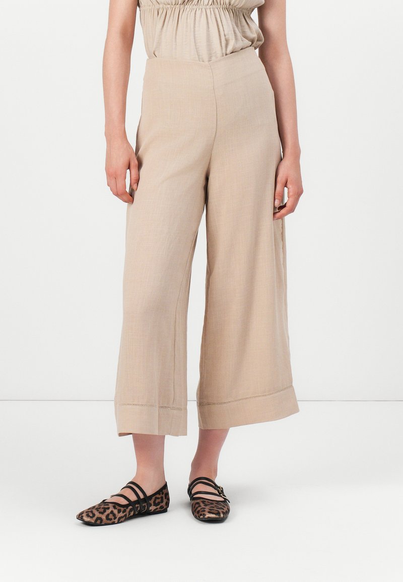 Pantalons beige à jambes larges avec une texture lisse, présentant une taille haute et des ourlets roulés, associés à des ballerines à imprimé léopard et un haut léger.