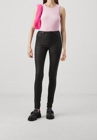 Jean skinny noir taille haute avec une finition lisse ; associé à un débardeur rose clair à côtes et à des chaussures noires chunky. Un sac fluffy rose est drapé sur l'épaule.