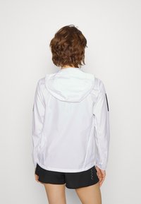 Valkoinen hupputakki, joka on valmistettu kiiltävästä ja kevyestä materiaalista. Sen hihoissa on mustia yksityiskohtia ja se on rento malli. Taaksepäin lyhyempi pituus.