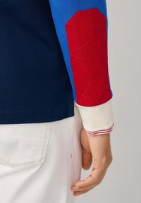 Person’s hand beside pants pocket wearing a blue shirt with a red padded elbow patch and white cuff with red stripes.