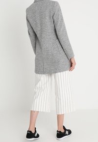 Grijze lange jas met notch-lapels, enkele achteropening, gecombineerd met witgestreepte culottes en zwarte sportschoenen. Soepele, gestructureerde stof.