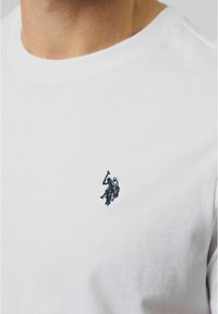 Balta medvilninė marškinėlių su apvaliu kaklu, kurios priekyje, kairiajame krūtinės šone, yra mažas tamsiai mėlynas išsiuvinėtas logotipas su raiteliu, laikantiu lazdą.