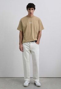 Oversized tričko s logem v béžové barvě, bílé džíny rovného střihu a bílé tenisky. Betonové pozadí, model stojí v neutrální póze.