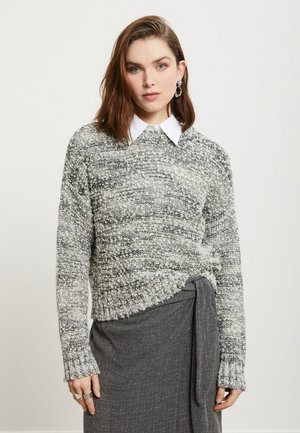 Jonge vrouw met bruin haar draagt een grijs-witte gestructureerde trui over een wit overhemd met kraag en een donkergrijze rok met zijstrik.