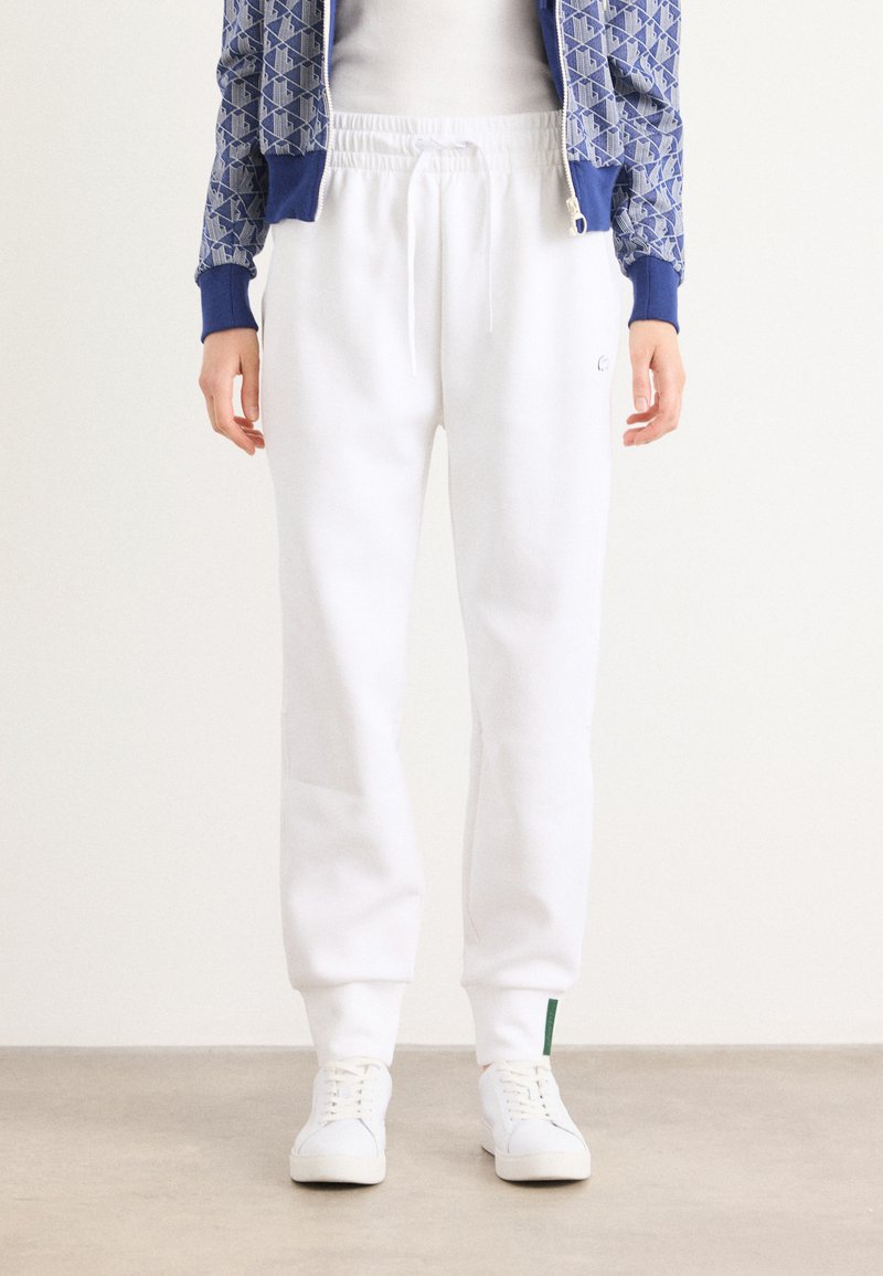 Vita sweatpants med dragsko i midjan och muddar vid anklarna, tillverkade av mjukt material. Har en grön detalj nära fållen.
