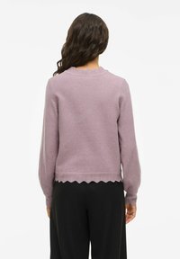 Femme avec de longs cheveux ondulés portant un pull lavande avec des bords festonnés et un pantalon noir, de dos contre un fond uni.