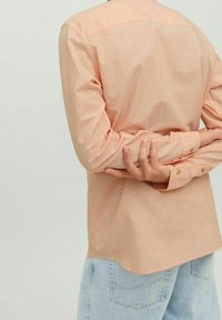 Camicia button-up color pesca chiaro con yoke sul retro, maniche lunghe e polsini. Indossata con jeans azzurri chiari, che mostrano una superficie liscia.
