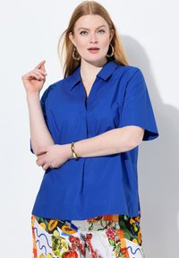 Camicia azzurra a maniche corte con colletto, caratterizzata da una piega frontale e una texture morbida. Abbinata a pantaloni colorati con motivo.