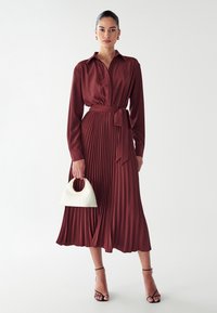 Pakaian plis marun dengan lengan panjang, kerah, dan ikat pinggang. Dilengkapi dengan tas tangan kecil berwarna putih dengan pegangan emas.