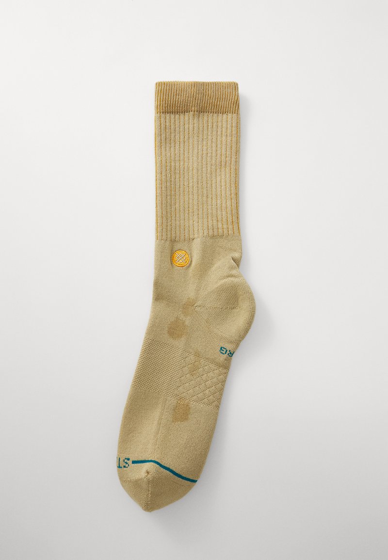 Beige sock with ribbed top, subtle patterned texture, logo patch in yellow, and turquoise accent near toe. Lightweight fabric, knee-high design.