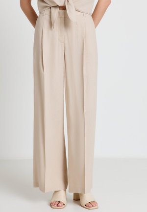 Pantalon beige taille haute à larges jambes, assorti à des chaussures à talons blocs ouvertes, porté avec un top beige noué.