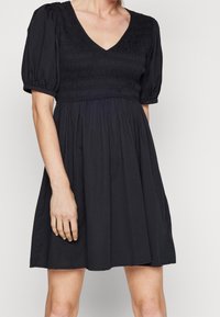 Svart klänning med V-ringning, korta puffärmar, smockad bodice och flödande kjol. Tillverkad av mjukt tyg med en slät yta.