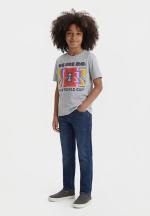 Chlapec s kučeravými vlasmi, ktorý má na sebe sivé tričko FIFA World Cup, modré rifle a sivé slip-on topánky, stojí s jednou rukou v vrecku.